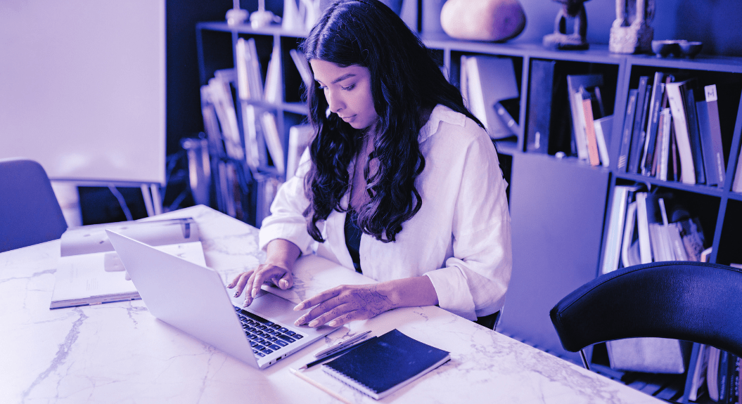 A woman with long hair focuses on UX design at a marble table in an office space. The background features a large bookshelf filled with various items. A notepad and pen rest on the table, complementing her work on crafting the ultimate guide to website layout. The image is tinted purple. | Ven Agency