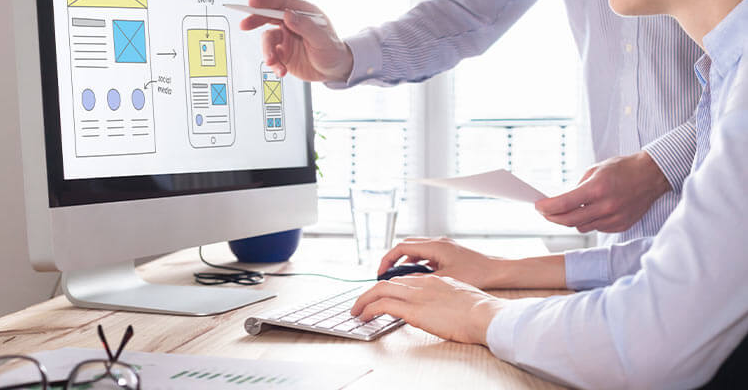 Two people collaborate on a web development project for a design agency. In the naturally lit Melbourne office, one points at wireframe designs on the monitor while the other types. Documents and a glass of water are scattered across the desk, as sunlight streams through the windows. | Ven Agency