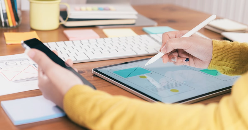 A person in a yellow sweater uses a stylus to design on a tablet displaying a web development wireframe. A smartphone is held in the other hand. On the wooden desk of this Melbourne web design agency, you'll find a keyboard, sticky notes, and a cup with pens. | Ven Agency