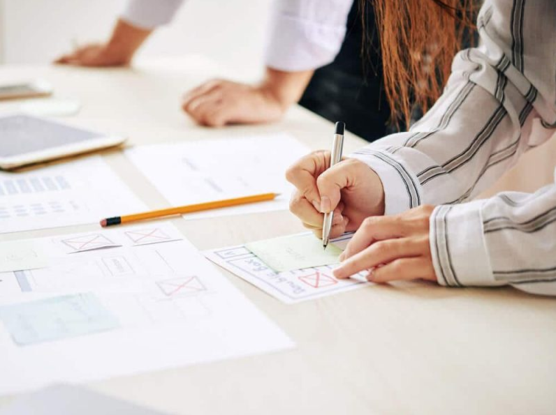 A person is jotting down ideas on sticky notes at a desk, surrounded by papers, a pencil, and a tablet. Another stands nearby with hands resting on the desk. The scene suggests a collaborative web design session in Melbourne. | Ven Agency