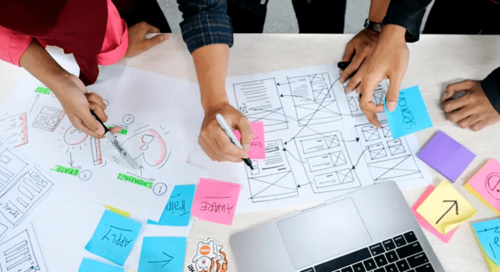 A group of people collaborate around a table covered with design sketches, sticky notes, and a laptop, diving into the ultimate guide for perfect website layout. Hands point and write on the papers, indicating an active UX design brainstorming session. | Ven Agency